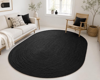 May include: A large, oval-shaped area rug featuring a solid black color and a braided, textured construction. The rug is positioned centrally on a light-colored floor, showcasing a dense, woven pattern that covers the entire surface area of the rug.