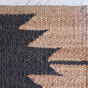May include: Close-up of a woven jute rug with a geometric design. The rug features a natural tan color with a bold black pattern. The edges are neatly finished, showcasing the rug's texture and craftsmanship.