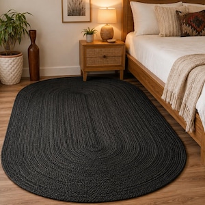 May include: An oval-shaped area rug featuring a dark charcoal black color and a braided texture. The rug is constructed from woven fibers arranged in concentric loops that follow the rounded perimeter of the piece, creating a uniform surface.