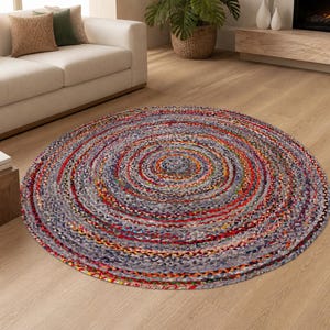Op de afbeelding: Een ronde gevlochten vloerkleed met een spiraalvormig ontwerp. Het vloerkleed heeft een mix van kleuren, waaronder grijs, rood, oranje en blauw. Het vloerkleed ligt op een lichtgekleurde houten vloer.