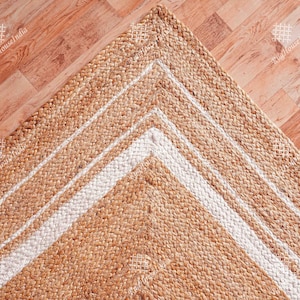 Puede incluir: Una alfombra de fibra natural con un borde blanco. La alfombra está hecha de yute y tiene una textura tejida. La alfombra es de color marrón claro y tiene un patrón geométrico.