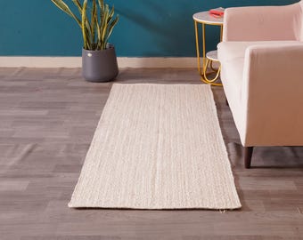 May include: A rectangular, off-white jute rug with a woven texture. The rug is placed on a wooden floor, with a pink sofa and a small side table in the background. A potted plant is also visible.