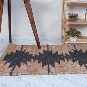 May include: A rectangular jute rug with a geometric black pattern. The rug is tan with a repeating zig-zag design in black. The rug is placed on a marble-patterned floor, with wooden table legs and a wooden shelf in the background.