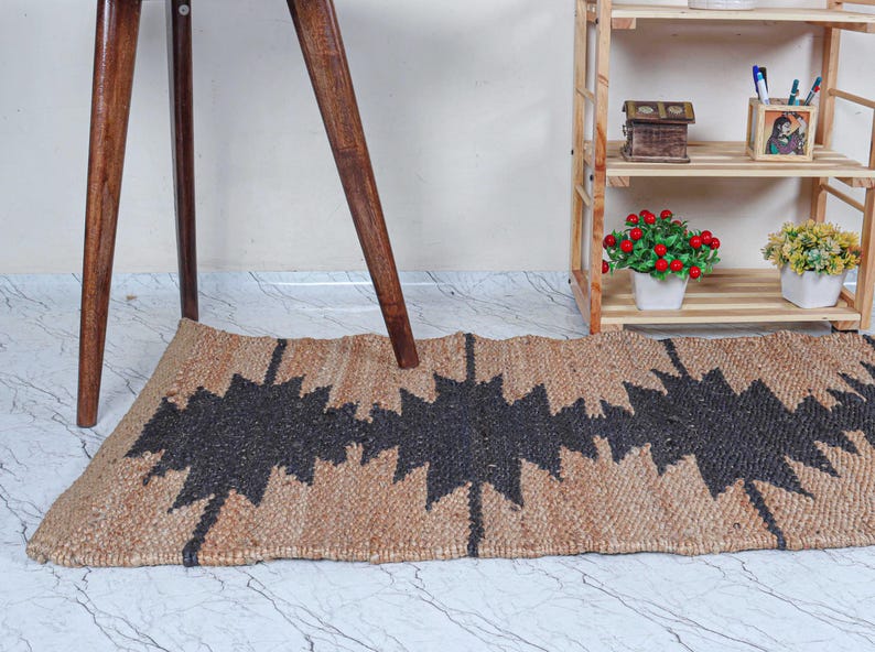 May include: A rectangular jute rug with a geometric black pattern. The rug is tan with a repeating zig-zag design in black. The rug is placed on a marble-patterned floor, with wooden table legs and a wooden shelf in the background.