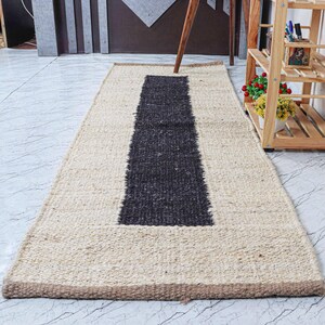 Op de afbeelding: Een rechthoekig jute vloerkleed met een centrale donkergrijze streep en een beige rand. Het vloerkleed is gemaakt van geweven vezels en heeft een textuur. Het vloerkleed ligt op een lichtgekleurde tegelvloer.