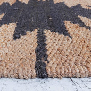 May include: Close-up of a natural jute rug with a black tree design. The rug has a woven texture with a mix of tan and black fibers. The tree design is centered and takes up a large portion of the rug.