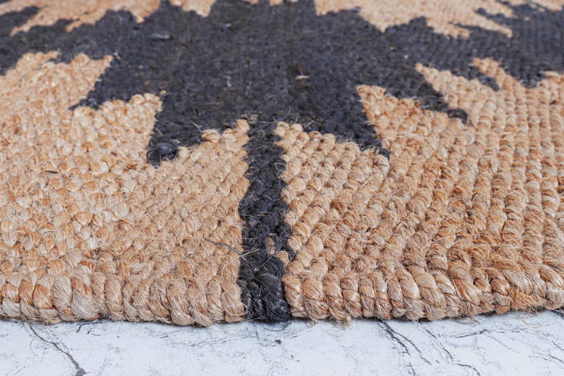 May include: Close-up of a natural jute rug with a black tree design. The rug has a woven texture with a mix of tan and black fibers. The tree design is centered and takes up a large portion of the rug.