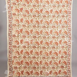 May include: Cream-coloured floral throw blanket with a repeating pattern of orange and green flowers and leaves. The blanket has a fringed edge and a border with a dark brown design. The overall design is symmetrical.