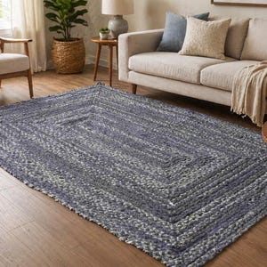 May include: A rectangular braided rug in shades of blue and white, with a geometric pattern. The rug is placed on a hardwood floor in a living room setting, with furniture visible in the background.