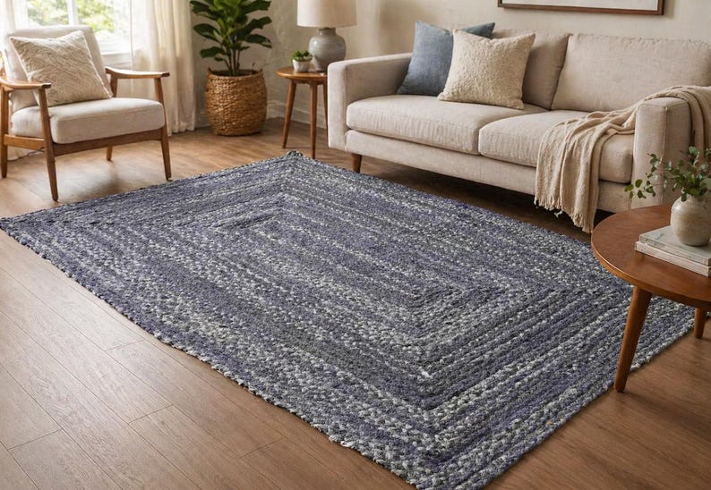 May include: A rectangular braided rug in shades of blue and white, with a geometric pattern. The rug is placed on a hardwood floor in a living room setting, with furniture visible in the background.