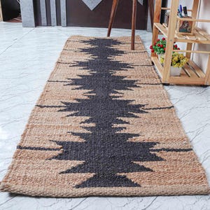 May include: A rectangular jute rug with a central dark gray geometric design. The rug has a natural tan border and a repeating pattern. The rug is placed on a white tile floor. The rug is approximately 60 inches long.