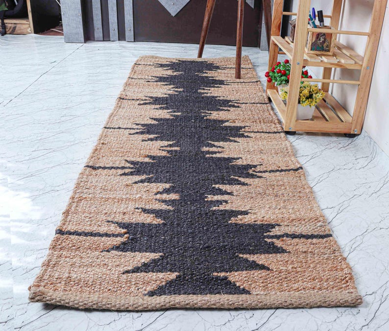 May include: A rectangular jute rug with a central dark gray geometric design. The rug has a natural tan border and a repeating pattern. The rug is placed on a white tile floor. The rug is approximately 60 inches long.