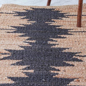 May include: A rectangular jute rug with a geometric design. The rug features a natural beige background with a central pattern in dark gray. The design is a series of zigzags, creating a textured, decorative floor covering.