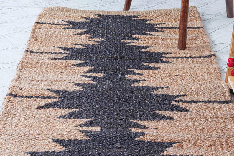 May include: A rectangular jute rug with a geometric design. The rug features a natural beige background with a central pattern in dark gray. The design is a series of zigzags, creating a textured, decorative floor covering.