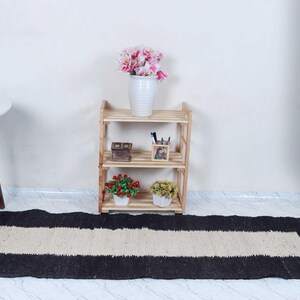 Op de afbeelding: Een rechthoekig zwart en beige tapijt met een centrale beige streep. Het tapijt ligt op een lichtgekleurde vloer. Achter het tapijt staat een houten plank met drie niveaus met decoratieve items en een witte vaas met roze bloemen.