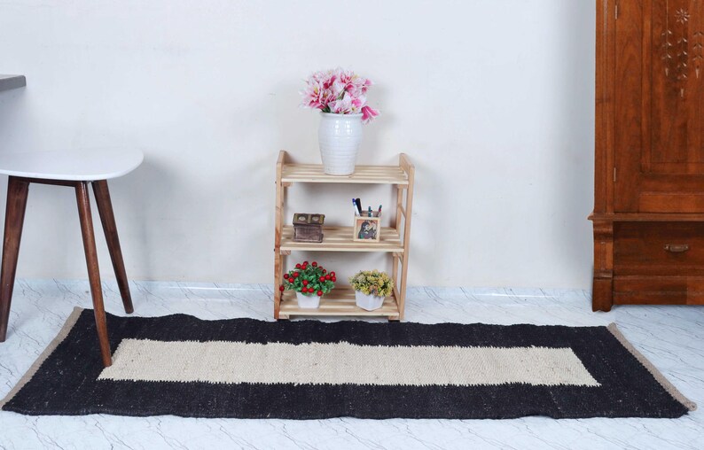 Op de afbeelding: Een rechthoekig zwart en beige tapijt met een centrale beige streep. Het tapijt ligt op een lichtgekleurde vloer. Achter het tapijt staat een houten plank met drie niveaus met decoratieve items en een witte vaas met roze bloemen.