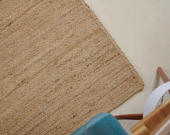 May include: A rectangular, woven jute rug in a natural tan color. The rug has a textured surface and is placed on a light-colored floor. A portion of a blue chair and a white chair are visible in the corner.