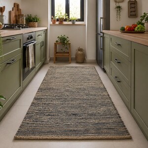 May include: A rectangular runner rug featuring a woven texture with alternating horizontal bands of dark navy blue and natural tan fibers. The rug is positioned centrally on a light-colored floor, extending lengthwise between two rows of olive green kitchen cabinets.