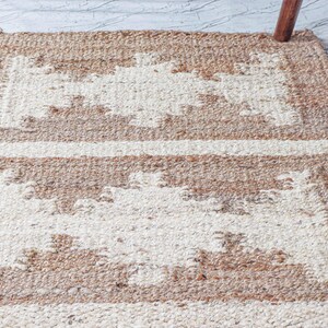 May include: A rectangular, woven rug with a geometric pattern. The rug features alternating bands of beige and off-white, creating a textured surface. The rug's design includes rectangular shapes and horizontal stripes, suitable for home decor.