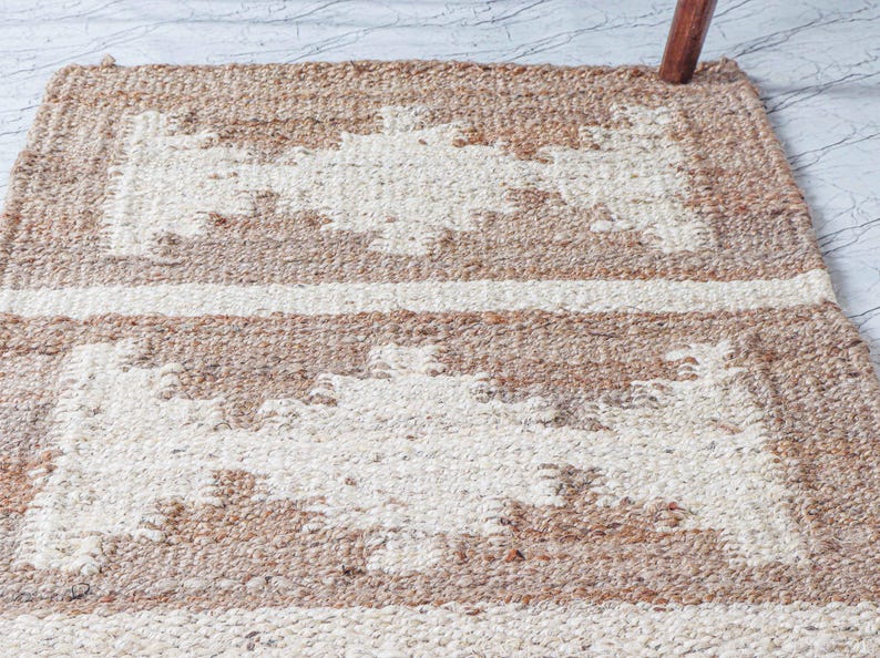 May include: A rectangular, woven rug with a geometric pattern. The rug features alternating bands of beige and off-white, creating a textured surface. The rug's design includes rectangular shapes and horizontal stripes, suitable for home decor.
