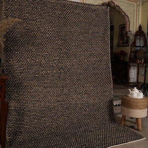 May include: A large, rectangular brown and beige woven rug with a textured pattern. The rug is displayed vertically, showcasing its size and design. The edges are finished with a white border. The rug is likely made of natural fibers.