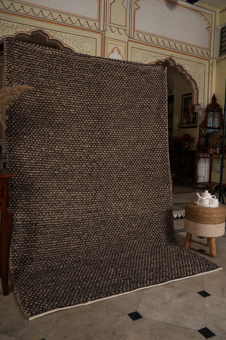 May include: A large, rectangular brown and beige woven rug with a textured pattern. The rug is displayed vertically, showcasing its size and design. The edges are finished with a white border. The rug is likely made of natural fibers.