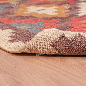 May include: Close-up of a brown, beige, and red patterned rug with a woven texture. The rug is rolled up and the edge is visible. The rug is on a wooden floor.