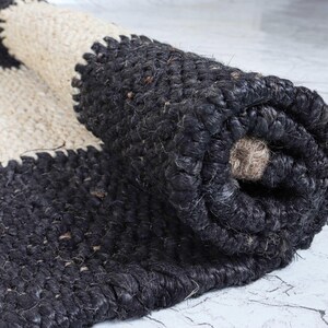 Op de afbeelding: Een opgerolde jute vloerkleed in zwart en cr&egrave;me. Het vloerkleed heeft een gestreept ontwerp met een getextureerde afwerking. Het zwarte gedeelte is dicht geweven, terwijl het cr&egrave;mekleurige gedeelte een lossere weving heeft. Het vloerkleed is gedeeltelijk uitgerold en toont de constructie.