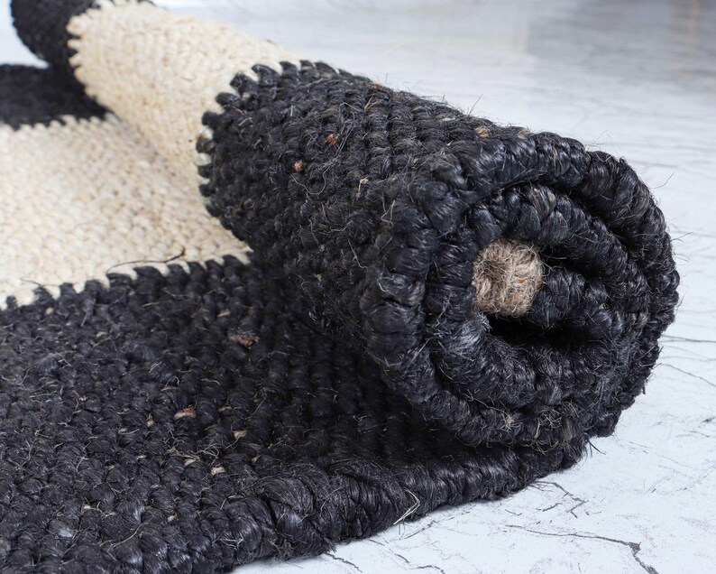 Op de afbeelding: Een opgerolde jute vloerkleed in zwart en cr&egrave;me. Het vloerkleed heeft een gestreept ontwerp met een getextureerde afwerking. Het zwarte gedeelte is dicht geweven, terwijl het cr&egrave;mekleurige gedeelte een lossere weving heeft. Het vloerkleed is gedeeltelijk uitgerold en toont de constructie.