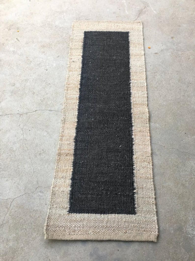 May include: A black and beige rectangular rug with a black center and a beige border. The rug is made of natural fibers and has a woven texture.
