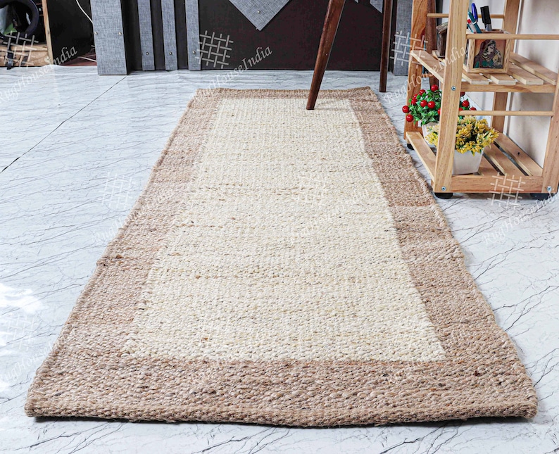 May include: A natural fiber rug with a light beige center and a brown border. The rug is made of woven jute and is perfect for adding a touch of natural texture to any room.