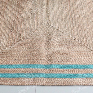 May include: A natural fiber rug with a woven design and a teal border. The rug has a square shape and is made of jute.