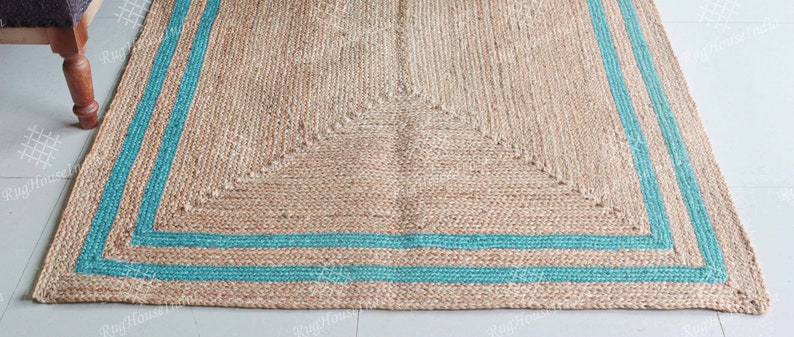 May include: A natural fiber rug with a woven design and a teal border. The rug has a square shape and is made of jute.