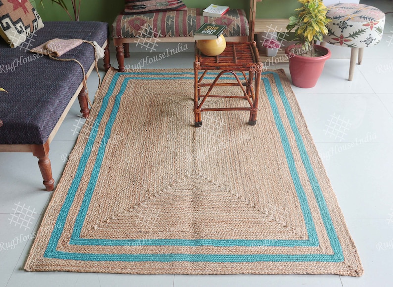 May include: A rectangular jute rug with a blue border. The rug has a woven pattern and is placed on a tiled floor. A small wooden table with a woven top sits on the rug. The table has a book and a yellow ceramic pot on top. A green plant in a red pot is in the background.