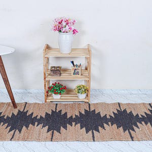 May include: A natural fiber runner rug with a geometric black pattern. The rug is placed on a light-colored floor. A small wooden shelf with decorative items and a white table with wooden legs are in the background.