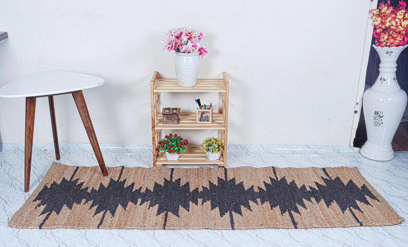 May include: A natural fiber runner rug with a geometric black pattern. The rug is placed on a light-colored floor. A small wooden shelf with decorative items and a white table with wooden legs are in the background.