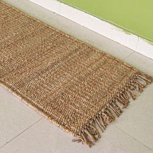 Pode incluir: Um tapete corredor retangular de juta tecida na cor marrom natural. O tapete tem uma borda com franjas em uma extremidade e é colocado em um piso de azulejos de cor clara. O tapete está inclinado para uma parede verde.