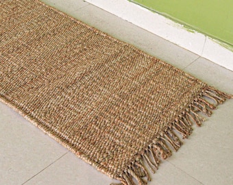May include: A rectangular, woven jute runner rug with a natural brown color. The rug has a fringed edge on one end and is placed on a light-colored tile floor. The rug is angled towards a green wall.