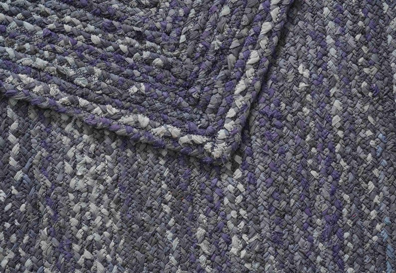 May include: Braided rug in shades of gray, white, and purple. The rug features a textured, woven design with a braided border. The color palette includes various shades of gray, with accents of white and purple.