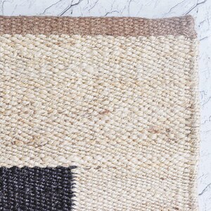 Op de afbeelding: Close-up van een geweven vloerkleed met een natuurlijke, getextureerde uitstraling. Het vloerkleed heeft een beige en bruin kleurenschema met een donker zwart vierkant patroon in de hoek. Het vloerkleed is gemaakt van een dik, duurzaam materiaal.