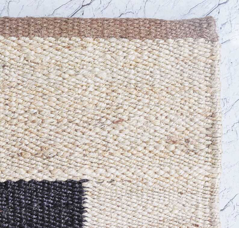 Op de afbeelding: Close-up van een geweven vloerkleed met een natuurlijke, getextureerde uitstraling. Het vloerkleed heeft een beige en bruin kleurenschema met een donker zwart vierkant patroon in de hoek. Het vloerkleed is gemaakt van een dik, duurzaam materiaal.