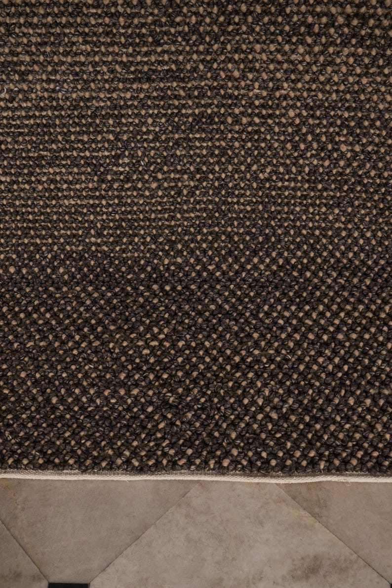 May include: Close-up of a textured, dark brown rug with a woven pattern. The rug has a neutral beige border and sits on a light brown tiled floor. The rug's surface is a mix of dark and light brown fibers.