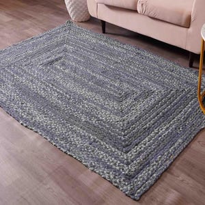 May include: A rectangular braided rug in shades of blue and gray, with a geometric pattern. The rug is placed on a wooden floor, with a sofa and side table in the background. The rug's texture is visible.