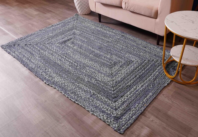 May include: A rectangular braided rug in shades of blue and gray, with a geometric pattern. The rug is placed on a wooden floor, with a sofa and side table in the background. The rug's texture is visible.