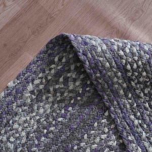 May include: Braided area rug in shades of gray, white, and purple. The rug has a textured appearance with a woven design. The corner of the rug is folded over, revealing the underside. The rug is on a wooden floor.