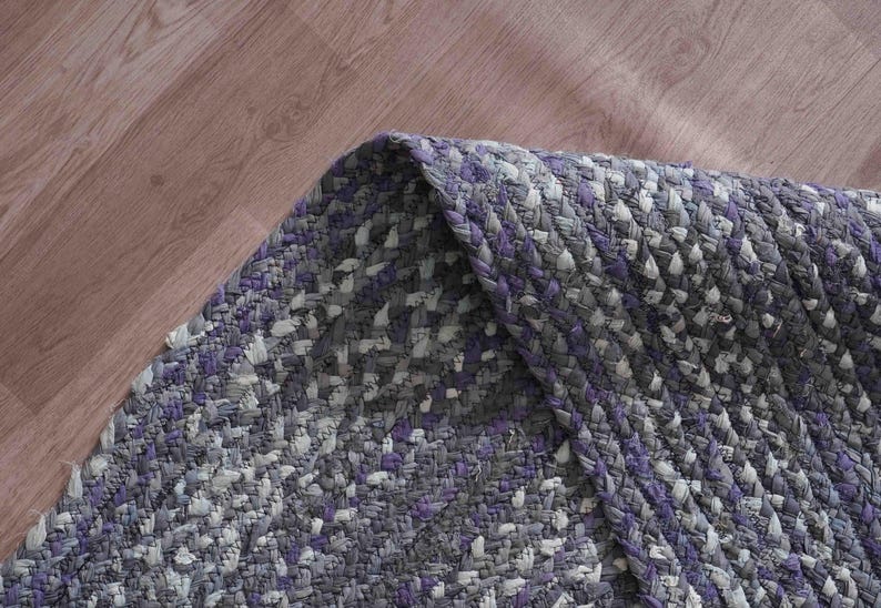 May include: Braided area rug in shades of gray, white, and purple. The rug has a textured appearance with a woven design. The corner of the rug is folded over, revealing the underside. The rug is on a wooden floor.
