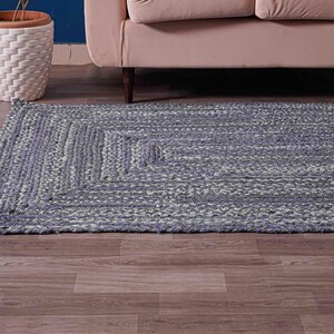 May include: A rectangular, braided rug in shades of blue and gray. The rug features a geometric pattern and is placed on a wooden floor. A white woven planter with a plant and a beige sofa are in the background.