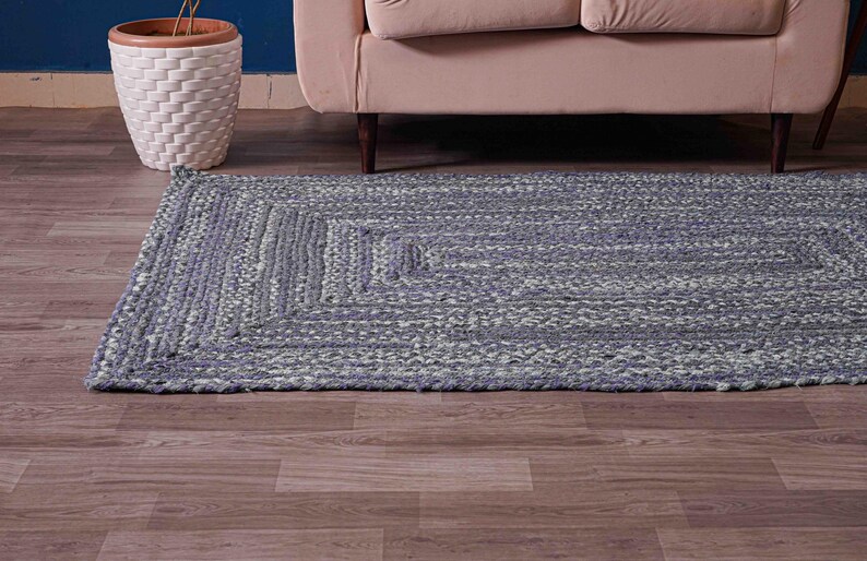 May include: A rectangular, braided rug in shades of blue and gray. The rug features a geometric pattern and is placed on a wooden floor. A white woven planter with a plant and a beige sofa are in the background.