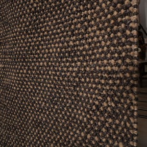 May include: Close-up of a textured rug with a repeating pattern. The rug features a combination of dark brown and tan colors, creating a visually interesting surface. The weave appears dense and durable, suitable for high-traffic areas.