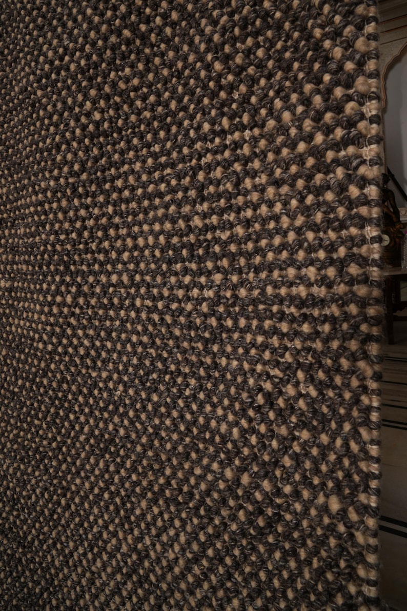 May include: Close-up of a textured rug with a repeating pattern. The rug features a combination of dark brown and tan colors, creating a visually interesting surface. The weave appears dense and durable, suitable for high-traffic areas.